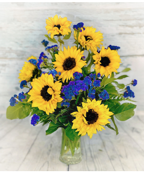 Midnight Sun are bright sunflowers, staggered in a tall style, accented with textured greenery with purple statis in a tall glass vase.